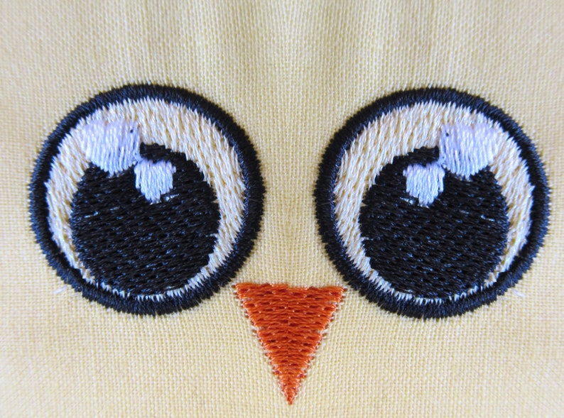 May include: Embroidered yellow fabric with two black eyes and an orange beak. The eyes are outlined in black and have white pupils.