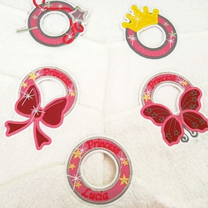Puede incluir: Cuatro anillos de toalla bordados con temas de princesa. Cada anillo presenta un diseño diferente: una varita mágica y una estrella, una corona, un lazo y una mariposa. Los anillos son todos de color rosa y gris con detalles dorados. La palabra "Princesa" está bordada en cada anillo.