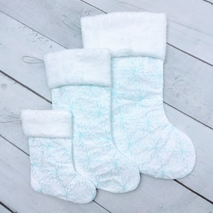 May include: Three white Christmas stockings with a snowflake pattern in light blue. The stockings are different sizes and have a fluffy white cuff.