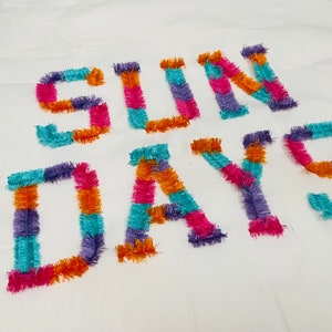 Peut inclure: Un tissu blanc avec les mots "SUN DAYS" brod&eacute;s en fil color&eacute;. Les lettres sont faites de fils de diff&eacute;rentes couleurs, dont l'orange, le rose, le violet et le turquoise.
