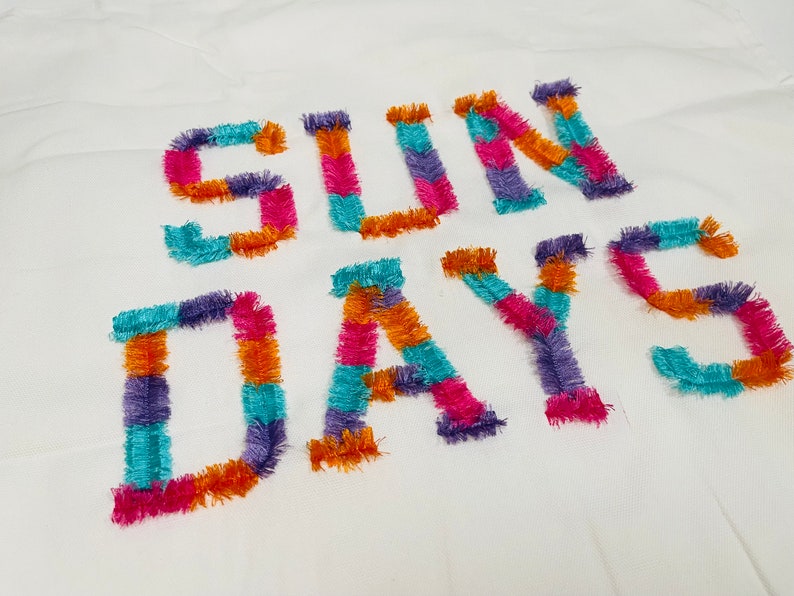 Peut inclure: Un tissu blanc avec les mots "SUN DAYS" brod&eacute;s en fil color&eacute;. Les lettres sont faites de fils de diff&eacute;rentes couleurs, dont l'orange, le rose, le violet et le turquoise.
