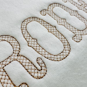 Può includere: Primo piano della parola "ROOM" ricamata in un carattere decorativo su un tessuto bianco. Le lettere sono delineate con un motivo marrone a punto croce, creando un effetto strutturato. Lo sfondo è un materiale bianco sporco.