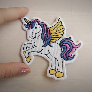 May include: Embroidered patch featuring a white unicorn with a yellow horn, wings, and hooves. The unicorn has a pink and blue mane and tail. The patch is outlined in white and is approximately 3 inches tall.