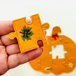 May include: A felt puzzle piece in the shape of a pumpkin, with a green and black embroidered leaf design. The piece is held in a hand, with the rest of the pumpkin puzzle visible in the background. The pumpkin is orange.