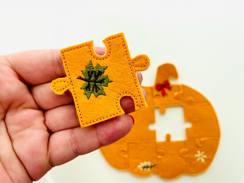 May include: A felt puzzle piece in the shape of a pumpkin, with a green and black embroidered leaf design. The piece is held in a hand, with the rest of the pumpkin puzzle visible in the background. The pumpkin is orange.