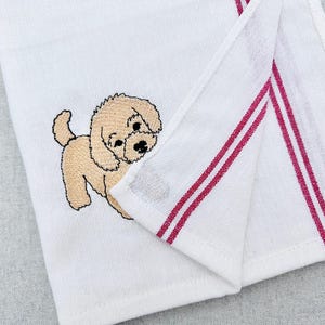 May include: White hand towel featuring an embroidered tan puppy. The towel has two red stripes along the edge. The puppy has a black nose and eyes. The towel is folded, displaying the embroidered puppy and the red stripes.