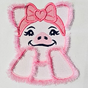 May include: Pink embroidered pig with a pink bow and a heart-shaped center. The pig has a smiling face with black eyes and a black nose.