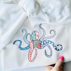 以下が含まれることがあります： カラフルな刺繍されたタコのデザインが施された白いスウェットシャツ。タコは、さまざまな模様の布の正方形でできています。