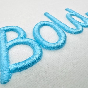 Puede incluir: Primer plano de la palabra "Bold" bordada en hilo azul claro sobre tela blanca. Las letras cursivas tienen una apariencia texturizada por la costura.