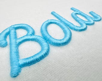 May include: Close-up of the word "Bold" embroidered in light blue thread on a white fabric. The letters are in a cursive style, with a textured appearance from the stitching.
