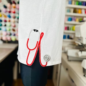 May include: White t-shirt with a red embroidered stethoscope design on the sleeve.