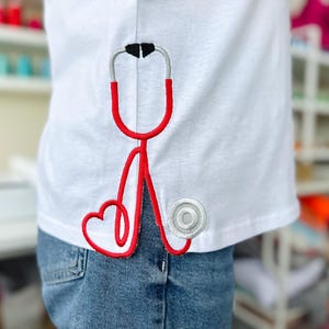 May include: White t-shirt with a red embroidered stethoscope design. The stethoscope has a heart shape at the bottom and a white circle on the side.