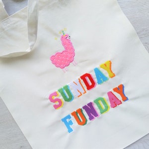 May include: A white canvas tote bag with the text "SUNDAY FUNDAY" embroidered in rainbow colors. A pink llama with a unicorn horn is embroidered above the text.