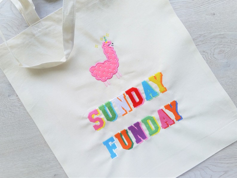 May include: A white canvas tote bag with the text "SUNDAY FUNDAY" embroidered in rainbow colors. A pink llama with a unicorn horn is embroidered above the text.