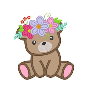 May include: A brown teddy bear with a floral crown. The bear has a light brown face and pink accents on its paws. The flower crown features pink, purple, and yellow flowers with green leaves. The bear is sitting with its legs extended.