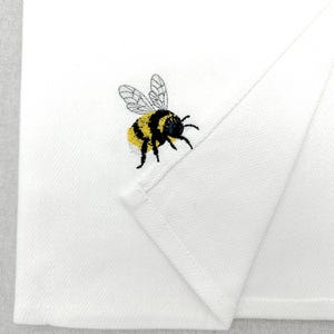 May include: A white kitchen towel featuring a small, embroidered bumblebee with yellow and black stripes and delicate, light grey wings. The fabric has a subtle herringbone weave pattern, and the towel is folded to show a clean, hemmed edge.