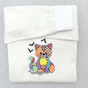 以下が含まれることがあります： 刺繍された猫のデザインが施された白い長袖シャツ。猫はジグザグ、チェック、ハートなど、さまざまなカラフルな幾何学模様で構成されています。猫の頭の上には、2匹の小さな黒いコウモリが刺繍されています。