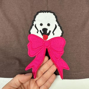 May include: White embroidered poodle with a pink bow on a brown fabric background.