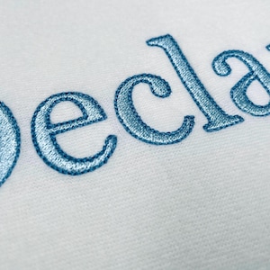 Puede incluir: Primer plano de tela blanca con el nombre "Declan" bordado en hilo azul claro. Las letras están en un estilo cursivo, con un ligero efecto de sombra, creando una apariencia texturizada.