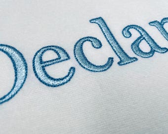 May include: Close-up of white fabric with the name "Declan" embroidered in light blue thread. The letters are in a cursive style, with a slight shadow effect, creating a textured appearance.
