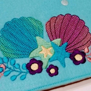Puede incluir: Parche de fieltro bordado con una concha azul y verde, una concha rosa, dos estrellas de mar azules y flores moradas con centros amarillos. El fondo es de color azul claro.