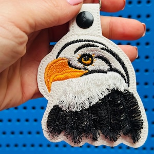 May include: White leather keychain with an embroidered eagle head. The eagle has a black and white feathered neck and a yellow eye.