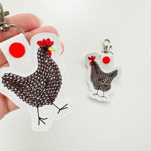 May include: Two chicken-shaped keyrings. The larger keyring is held by a hand. Both keyrings feature a white background with a brown embroidered chicken design, a red comb, and a red circle. The keyrings have silver-coloured clasps.