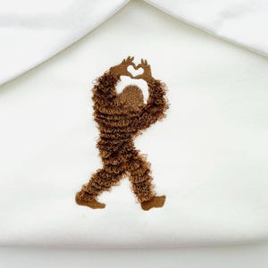 May include: White fabric with a brown embroidered design of a Bigfoot figure. The Bigfoot is walking and forming a heart shape with its hands. The design is centered on the fabric.