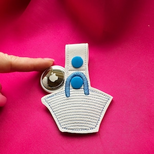 Op de afbeelding: Wit leren AirPod-houder met blauwe stiksels en twee blauwe knoppen. De houder is gevormd als een mand en heeft een wit en blauw gestreept patroon.