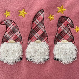 May include: Three gnomes with red and black plaid hats and white fuzzy beards. The gnomes are embroidered on a pink fabric background with yellow stars.