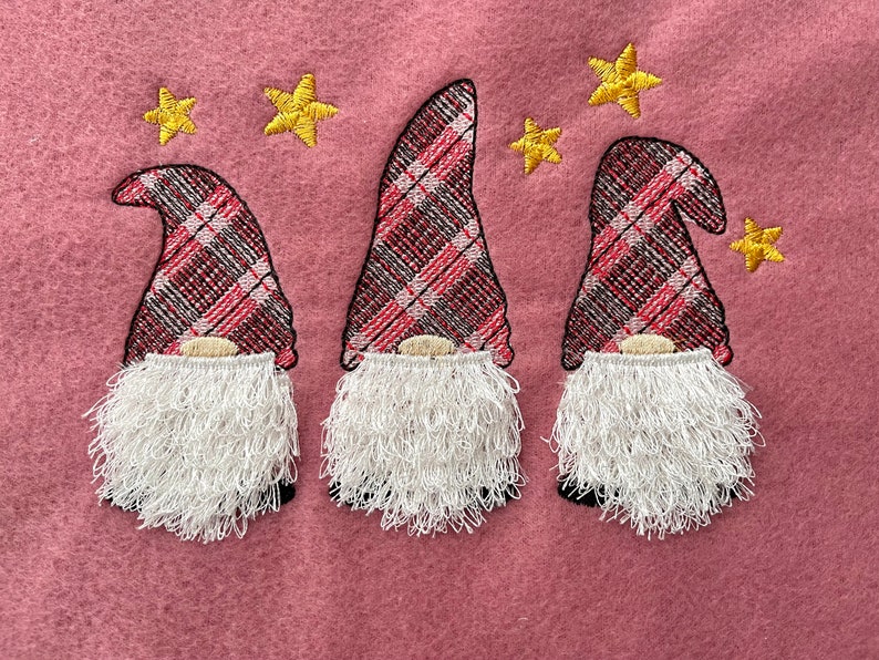 May include: Three gnomes with red and black plaid hats and white fuzzy beards. The gnomes are embroidered on a pink fabric background with yellow stars.