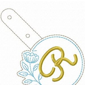 May include: A gold embroidered letter "R" with a blue floral design in a circle. The circle is attached to a gray strap.
