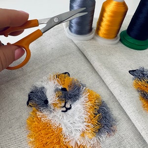 May include: Close-up of a fluffy, embroidered animal with yellow, white, and gray fur. The animal is stitched onto a light gray fabric. A pair of orange handled scissors are in the foreground.