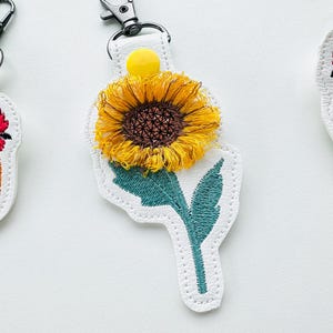 May include: White leather keychain with a colorful embroidered sunflower design. The sunflower has yellow petals, a brown center, and green leaves. The keychain has a metal ring.