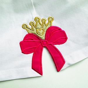 Puede incluir: Una tela blanca con un lazo rosa y una corona dorada con un corazón en el centro.