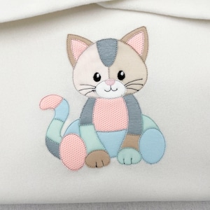 May include: A white fabric surface featuring a multi-colored cat appliqué. The cat is composed of various pastel-colored fabric patches in shades of pink, blue, grey, and tan, with black embroidered outlines, whiskers, eyes, and a small nose.