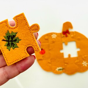 May include: A felt puzzle piece in the shape of a pumpkin, with a matching piece held in hand. The puzzle pieces are orange with green and white embroidered details. The pumpkin puzzle has a bow and snowflake details.