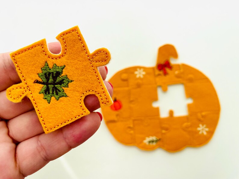 May include: A felt puzzle piece in the shape of a pumpkin, with a matching piece held in hand. The puzzle pieces are orange with green and white embroidered details. The pumpkin puzzle has a bow and snowflake details.