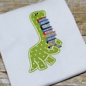 May include: Green dinosaur with white polka dots carrying a stack of colourful books. The dinosaur is smiling and has a green outline.