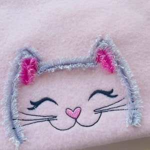 May include: Embroidered gray cat face with pink ears and a pink heart nose on a pink fleece fabric.