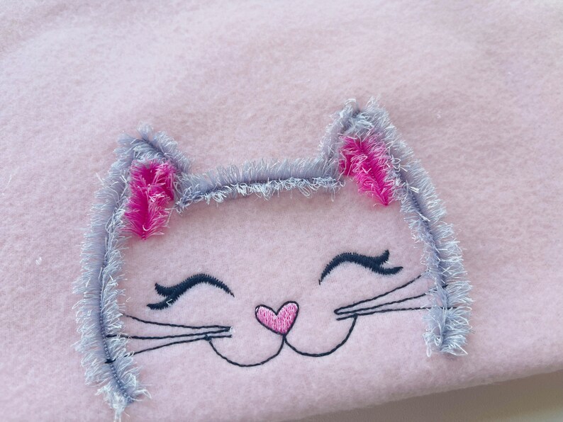 May include: Embroidered gray cat face with pink ears and a pink heart nose on a pink fleece fabric.