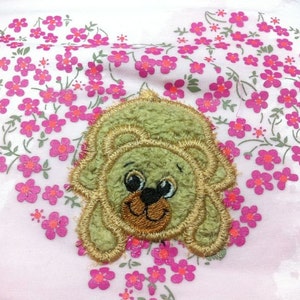 May include: A green plush bear with brown eyes and a brown nose is embroidered on a pink fabric background with a pattern of pink flowers.