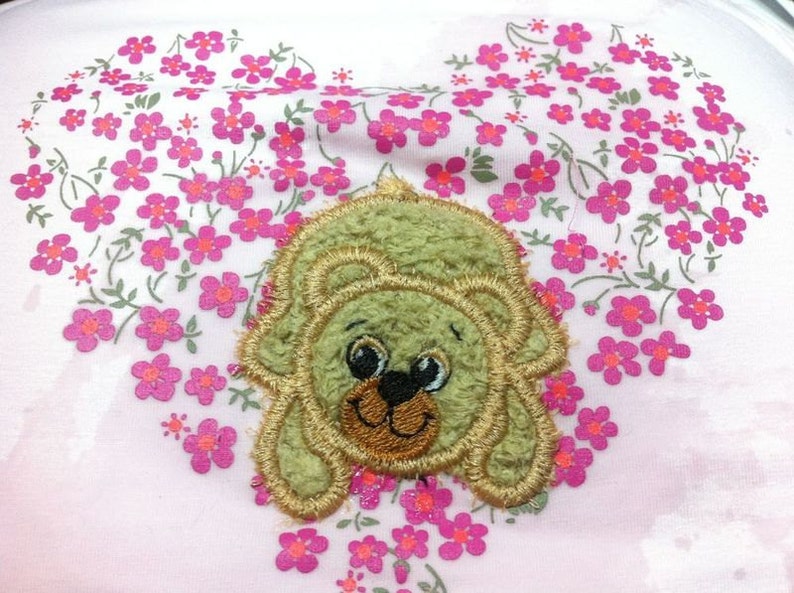 May include: A green plush bear with brown eyes and a brown nose is embroidered on a pink fabric background with a pattern of pink flowers.
