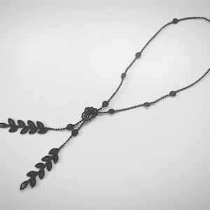 Può includere: Una collana nera con una lunga catena e due ciondoli a forma di foglia. La catena è fatta di piccole perle nere.