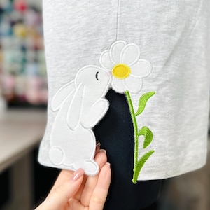 以下が含まれることがあります： グレーのシャツに白いウサギが刺繍されており、黄色の芯を持つ白いデイジーの花を嗅いでいます。デイジーは緑の葉のついた茎に付いています。
