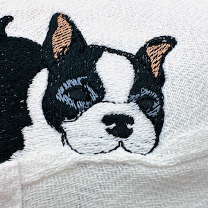 May include: Embroidered Boston Terrier dog on a white fabric. The dog's face is black and white with tan ears. The embroidery is detailed, showing the dog's features. The fabric has a woven texture.