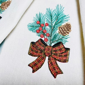 Op de afbeelding: Geborduurde keukendoek met een groene dennentak, rode bessen, een bruine dennenappel en een rood-gouden strik.