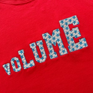 May include: Red t-shirt with the word "VOLUME" embroidered in white with blue star accents.