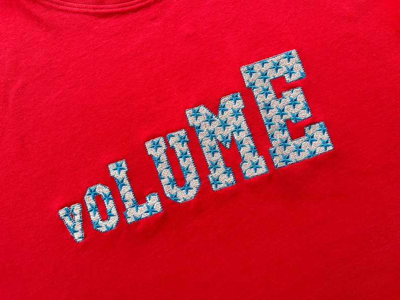 May include: Red t-shirt with the word "VOLUME" embroidered in white with blue star accents.