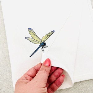 May include: A white fabric towel featuring an embroidered dragonfly with yellow wings and a dark blue body. The dragonfly is positioned on the upper portion of the towel, which is folded to reveal the texture of the white fabric material.